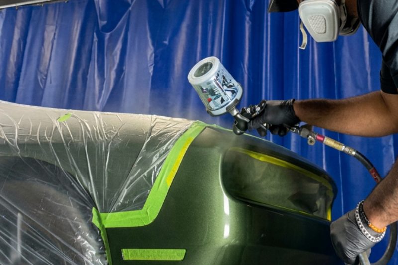 A technician spraying paint onto a masked green bumper inside the paint booth